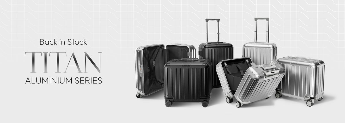 A promotional graphic for the Titan Aluminium Series luggage, showing multiple hard-shell suitcases in black and silver aluminum finishes, some standing upright and others open to show the interior. The text 'Back in Stock' and 'TITAN ALUMINIUM SERIES' is displayed on the left side against a light gray background with a subtle grid pattern.